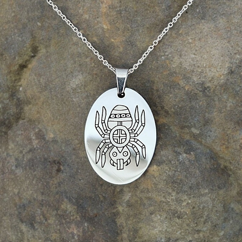 May include: A silver chain necklace with an oval pendant featuring a detailed engraved design of a spider.