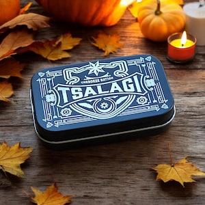 May include: A black metal tin with a white design featuring the words "Cherokee Nation" and "TsalaGi". The tin is surrounded by fall leaves on a wooden surface.