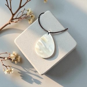 May include: A pearlescent white pendant necklace with a teardrop-shaped shell pendant. The pendant hangs from a brown cord and is set against a white square surface. Delicate branches with small white flowers are in the background.