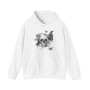 May include: White hoodie featuring a graphic print of a skull surrounded by flowers, leaves, and birds. The skull is white with black eyes and a black nose. The flowers are in shades of pink, purple, and yellow. The leaves are green and brown. The birds are black and white.