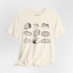 Op de afbeelding: Een crèmekleurig T-shirt met een zwart-witte illustratie van verschillende soorten brood. De tekst "My Shop Inc." is op het shirt gedrukt.