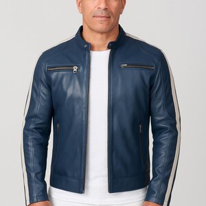 Puede incluir: Chaqueta de cuero azul con cuello alzado y cierre de cremallera. La chaqueta presenta dos rayas verticales color crema en las mangas y dos bolsillos en el pecho con cremallera. El modelo lleva una camiseta blanca y pantalones blancos.