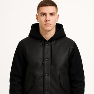 May include: Black hooded bomber jacket with a leather-look front panel and black sleeves. The jacket features a button-up closure, two front pockets, and a ribbed hem and cuffs. The model is wearing black jeans.