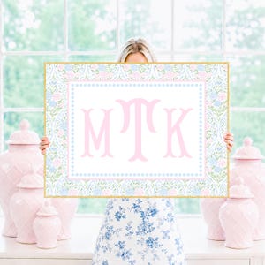 May include: A rectangular sign with a gold frame featuring a white center with the large pink monogram letters MTK. The border displays a repeating pattern of light blue and pink flowers with green leaves, enclosed by a light blue dotted line.