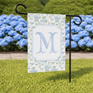 May include: A garden flag featuring a light blue letter "M" within a white and light blue frame. The flag has a floral pattern with blue hydrangeas and is displayed on a black metal stand in a garden setting.