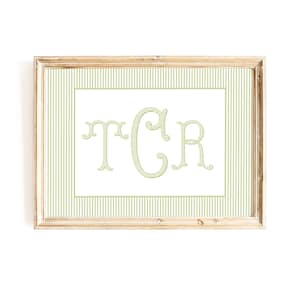 Könnte beinhalten: Ein rechteckiger gerahmter Druck mit einem weißen Mittelfeld und den großen, stilisierten grünen Monogrammbuchstaben TCR. Der Rand besteht aus einem vertikalen grün-weißen Nadelstreifenmuster. Der Rahmen ist aus hellem Holz mit natürlichem Finish.