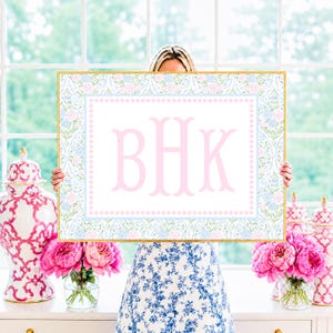 Op de afbeelding: Een rechthoekig bord met de letters BHK in een groot roze schreeflettertype, gecentreerd in een wit veld. De rand bestaat uit een lichtblauwe stippellijn omringd door een bloemenpatroon van roze en blauwe bloemen op een witte achtergrond.