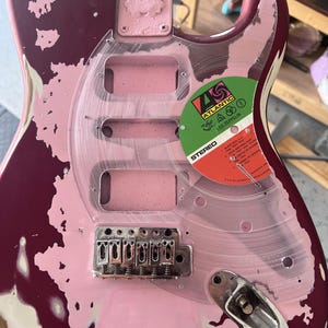 Puede incluir: Guitarra el&eacute;ctrica parcialmente desmontada con acabado rosa y burdeos. Se ve un golpeador de pl&aacute;stico transparente, junto con el puente y una etiqueta de disco de Led Zeppelin. La guitarra muestra signos de desgaste y reparaci&oacute;n.