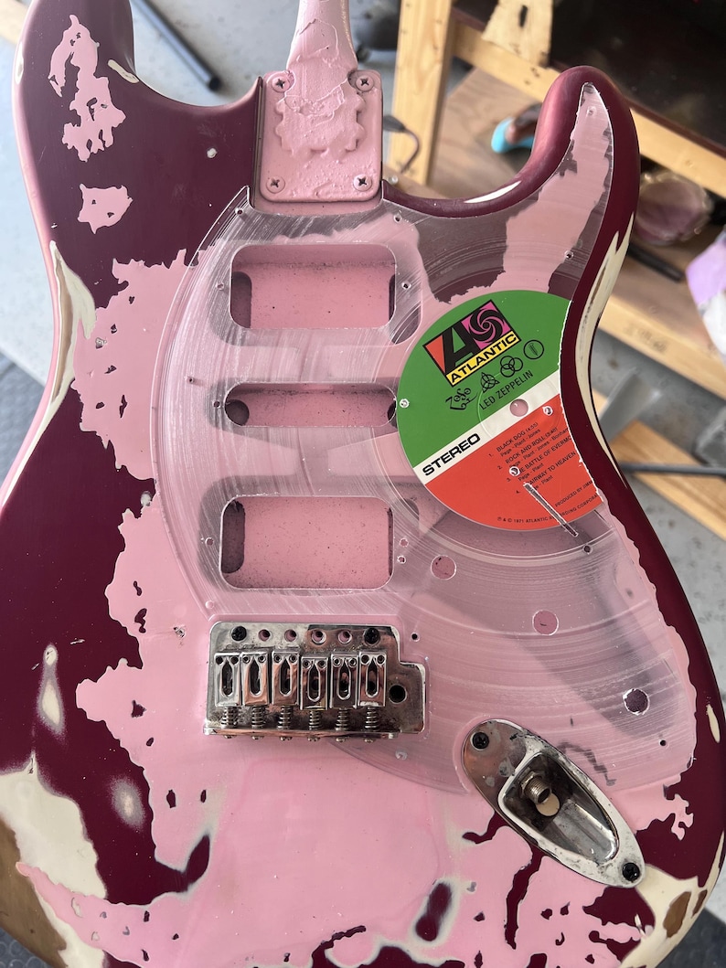 Puede incluir: Guitarra el&eacute;ctrica parcialmente desmontada con acabado rosa y burdeos. Se ve un golpeador de pl&aacute;stico transparente, junto con el puente y una etiqueta de disco de Led Zeppelin. La guitarra muestra signos de desgaste y reparaci&oacute;n.