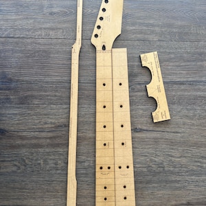 Puede incluir: Un conjunto de plantillas para mástil de guitarra hechas de madera marrón claro. El conjunto incluye la pala, el diapasón y otros componentes. Las plantillas están dispuestas sobre una superficie de madera oscura. La pala tiene agujeros pre-perforados para las clavijas.