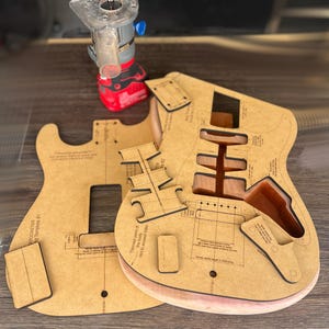 Pode incluir: Um projeto de marcenaria com modelos de corpo de guitarra e uma roteadora vermelha e prateada. Os modelos são feitos de madeira marrom clara e têm marcações detalhadas. A roteadora está posicionada acima dos modelos.