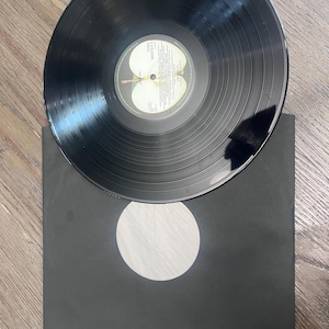 Puede incluir: Un disco de vinilo negro con una etiqueta blanca y amarilla con texto y un logotipo de manzana verde. El disco est&aacute; colocado encima de una funda de papel negra con un recorte circular blanco.
