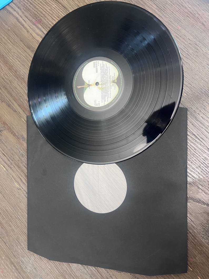 Puede incluir: Un disco de vinilo negro con una etiqueta blanca y amarilla con texto y un logotipo de manzana verde. El disco est&aacute; colocado encima de una funda de papel negra con un recorte circular blanco.