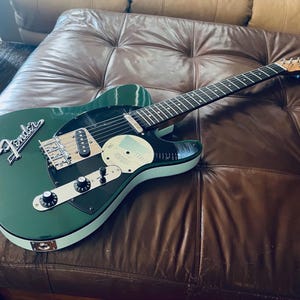 Puede incluir: Una guitarra el&eacute;ctrica Fender Telecaster verde oscuro con herrajes cromados. La guitarra presenta un golpeador negro, una placa de control blanca y una pegatina redonda en el cuerpo. El logotipo de Fender es visible en el clavijero.