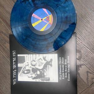 Puede incluir: Un disco de vinilo azul y negro veteado sobre una funda de &aacute;lbum negra. La funda presenta una foto en blanco y negro de una banda y el texto "OZZY OSBOURNE DIARY OF A MADMAN".