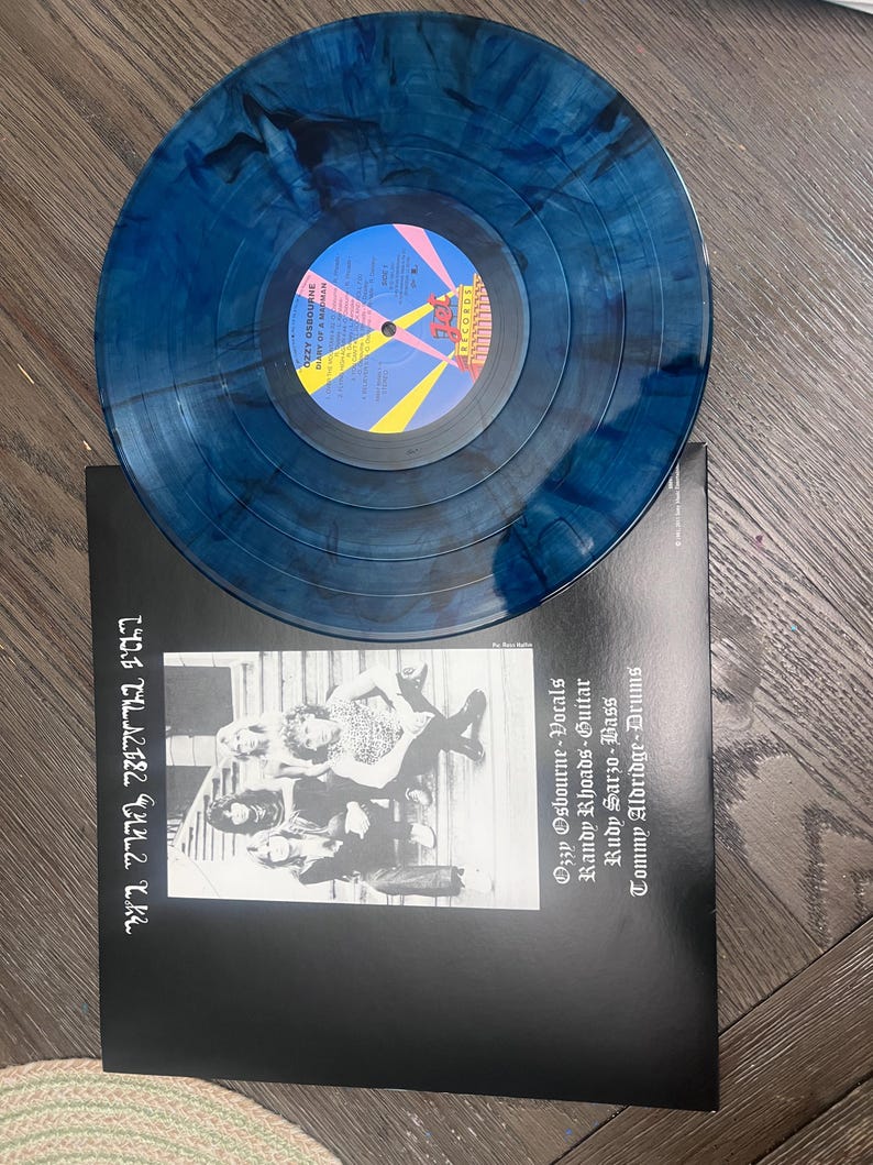 Puede incluir: Un disco de vinilo azul y negro veteado sobre una funda de &aacute;lbum negra. La funda presenta una foto en blanco y negro de una banda y el texto "OZZY OSBOURNE DIARY OF A MADMAN".