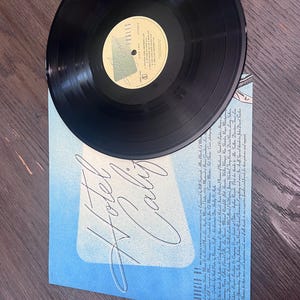 Puede incluir: Un disco de vinilo negro sobre una portada de &aacute;lbum azul. La portada del &aacute;lbum tiene las palabras "Hotel California" en una escritura estilizada. La etiqueta del disco es visible, con texto y un logotipo. El &aacute;lbum est&aacute; sobre una superficie de madera.