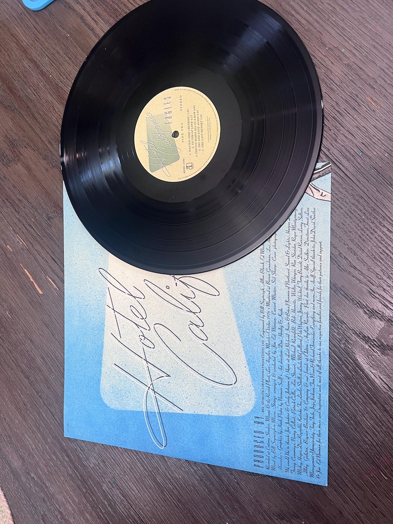 Puede incluir: Un disco de vinilo negro sobre una portada de &aacute;lbum azul. La portada del &aacute;lbum tiene las palabras "Hotel California" en una escritura estilizada. La etiqueta del disco es visible, con texto y un logotipo. El &aacute;lbum est&aacute; sobre una superficie de madera.