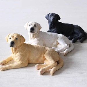 May include: Three Labrador Retriever figurines in yellow, white, and black. Each dog is lying down, with front paws extended. The figurines have a smooth finish and are detailed to resemble the breed. The dogs are arranged in a row.