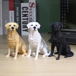 Puede incluir: Tres figuras de Labrador Retriever. Se muestra uno amarillo, uno blanco con manchas grises y uno negro. Cada perro está sentado erguido. Las figuras están sobre una superficie marrón claro.