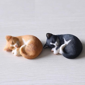 May include: Two small ceramic cat figurines, one orange and white and one black and white, are curled up asleep. The cats are both facing the right side of the image.