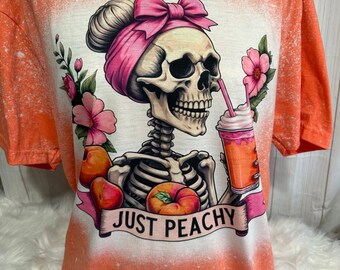 May include: Orange bleached t-shirt with a graphic of a skeleton wearing a pink headband and bow, holding a peach smoothie with a straw. The text "JUST PEACHY" is printed below the skeleton.