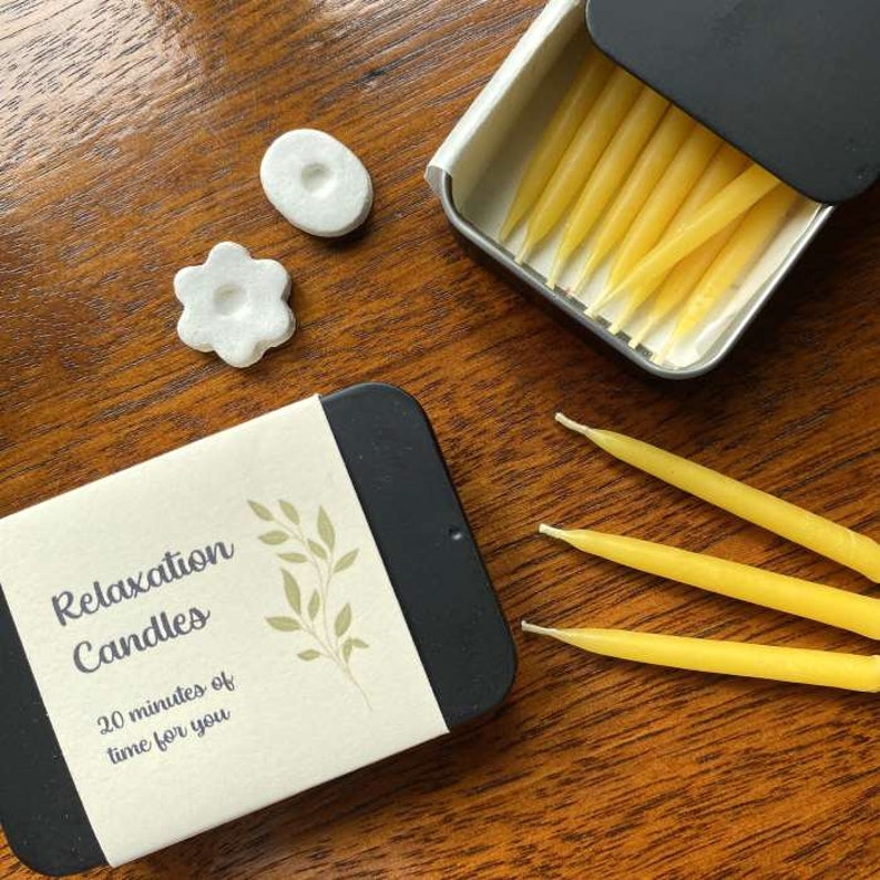 Two tins of Relaxation Candles, one open to show the beeswax mini taper candles inside, with three 20 minutes candles and two candle holders lying on the table next to them