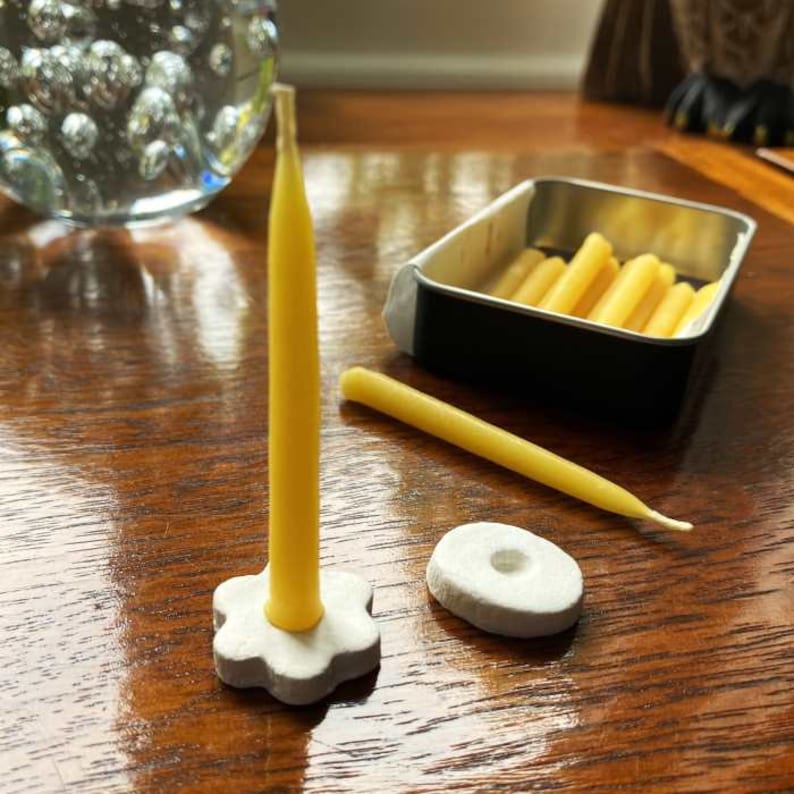 A 20 minute candle made of pure beeswax standing in a simple candle holder with a second mini taper candle lying behind it and with an open tin of Relaxation Candles in the background