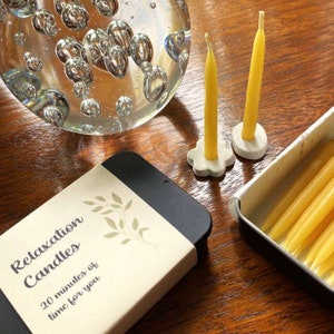 Two beeswax mini taper candles standing in simple ceramic holders on a wooden table, with two tins of Relaxation Candles in front, one open to show the beeswax candles inside and one closed.