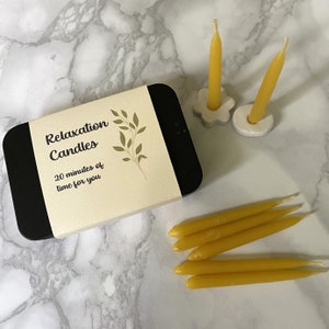 Black tin of relaxation candles on a marble counter with two 20 minute candles standing in holders and further beeswax mini taper candles lying nearby