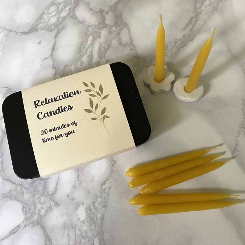 Black tin of relaxation candles on a marble counter with two 20 minute candles standing in holders and further beeswax mini taper candles lying nearby