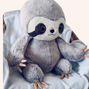 Pode incluir: Uma pelúcia de bicho-preguiça com pelo cinza macio, rosto cor de creme e manchas marrom-escuras ao redor dos olhos. O brinquedo de pelúcia apresenta um pequeno nariz preto, boca costurada e garras cor de bege nas mãos e nos pés.