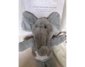 May include: A grey plush elephant pacifier holder attached to a cardboard backing card. The card features the text Keep me Close PLUSH PACIFIER HOLDER. The elephant has large ears, a trunk, and a gold-coloured band around its midsection.
