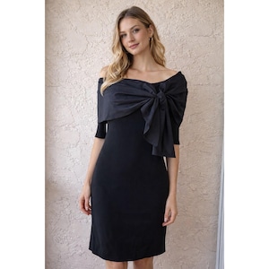 May include: Black off-the-shoulder dress with a large bow detail. The dress has short sleeves and a knee-length hemline. The bow is made of a shiny fabric, creating a striking contrast with the matte finish of the dress. A classic and elegant design.