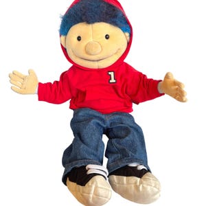 May include: A plush doll of a boy wearing a red hooded sweatshirt with the number 1, blue jeans, and black and white sneakers. The doll has blue hair and a smiling face with open arms.