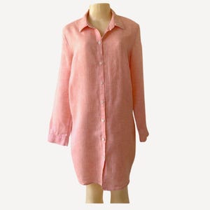 May include: A long-sleeved button-down shirt in a soft coral-pink hue. The garment features a classic collar, a full-length front button placket with matching light-colored buttons, and a straight hemline, crafted from a textured fabric.