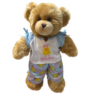 May include: A plush brown teddy bear wearing a white and light blue shirt with small blue bows on the shoulders and a yellow chick graphic wearing a pink crown with the text "CHICKS RULE". The bear wears matching light blue patterned shorts.