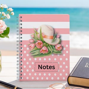 Puede incluir: Un cuaderno en espiral rosa con un diseño de sombrero y tulipanes, y la palabra «Notes». La portada tiene un estampado de lunares y rayas. También se ven un bolígrafo, gafas y un libro. En el fondo se ve el mar.