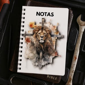 May include: A spiral-bound notebook with the word "NOTAS" on the cover, featuring a watercolour illustration of a lion superimposed on a cross. The notebook rests on a Bible, with tools in the background.