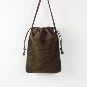 Pode incluir: Uma bolsa de couro marrom com cordão de ajuste e corpo de tecido tweed verde e marrom. A bolsa tem uma alça longa de couro marrom.