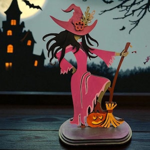 May include: A pink witch figure with a matching hat and dress, holding a broom. A carved pumpkin sits at the base. The background features a spooky castle, bats, and a full moon, creating a Halloween theme.