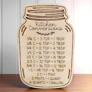 May include: Wooden kitchen conversion chart in the shape of a mason jar. The text "Kitchen Conversions" is at the top, with measurements for cups, teaspoons, tablespoons, pints, quarts, and gallons. The chart is made of light-colored wood.