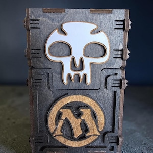 May include: A dark brown wooden box with a rectangular shape and interlocking joints. The front features a white skull cutout at the top and a circular wooden emblem with the letter M in the center, showcasing a layered, laser-cut design.