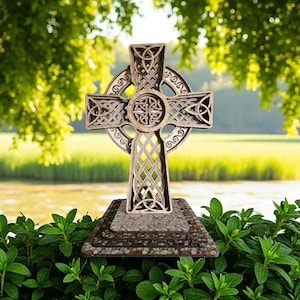 Może przedstawiać: Drewniany krzyż celtycki z misternym wzorem węzłów, stojący na kamiennej podstawie. Krzyż jest w jasnobrązowym kolorze, ze szczegółowym wzorem. Tło przedstawia zieleń i wodę.