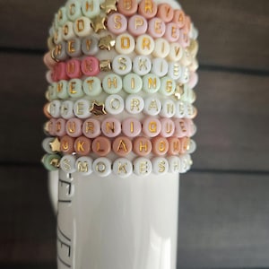 May include: A stack of colorful beaded bracelets with gold letter charms spelling out words like "OKLAHOMA", "SMOKESH", "UNIQUE", "ORANGE", "SOMETHING", "SKIES", "DRIVE", "SPEED", "BRYAN", and "GOODS".
