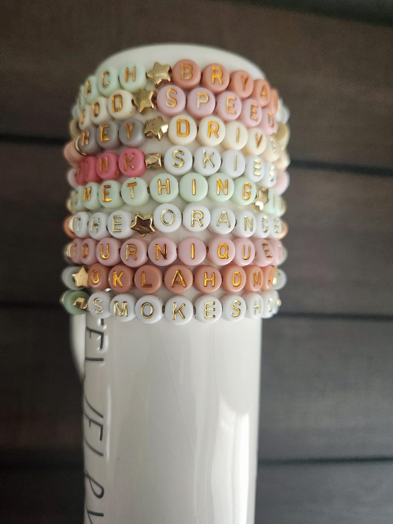 May include: A stack of colorful beaded bracelets with gold letter charms spelling out words like "OKLAHOMA", "SMOKESH", "UNIQUE", "ORANGE", "SOMETHING", "SKIES", "DRIVE", "SPEED", "BRYAN", and "GOODS".