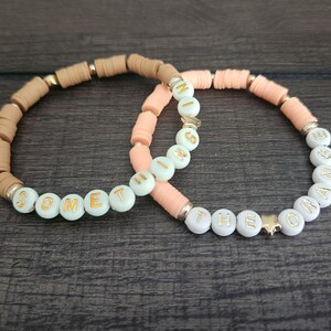 May include: Two beaded bracelets with the words "Something" and "The Orange" spelled out in gold letters on white beads. The bracelets are made with peach and brown beads.