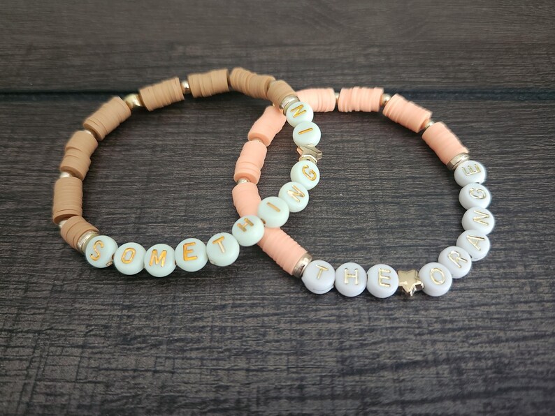 May include: Two beaded bracelets with the words "Something" and "The Orange" spelled out in gold letters on white beads. The bracelets are made with peach and brown beads.