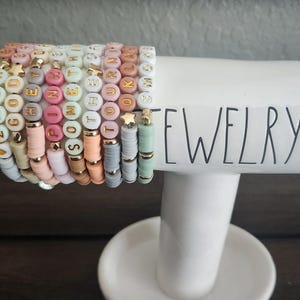 May include: A stack of colorful beaded bracelets with gold letter charms spelling out words like "ZACHARY", "PINK", and "SOMETHING". The bracelets are displayed on a white ceramic jewelry stand with the word "JEWELRY" printed on it.