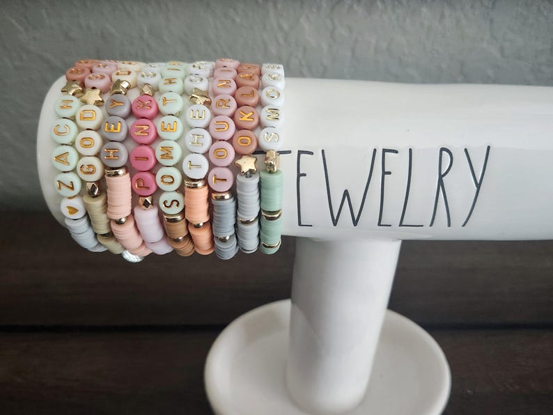 May include: A stack of colorful beaded bracelets with gold letter charms spelling out words like "ZACHARY", "PINK", and "SOMETHING". The bracelets are displayed on a white ceramic jewelry stand with the word "JEWELRY" printed on it.